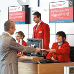 An Austrian Airlines baggage drop-off desk at Vienna Airport Terminal 3 with staff in red uniforms serving a passenger