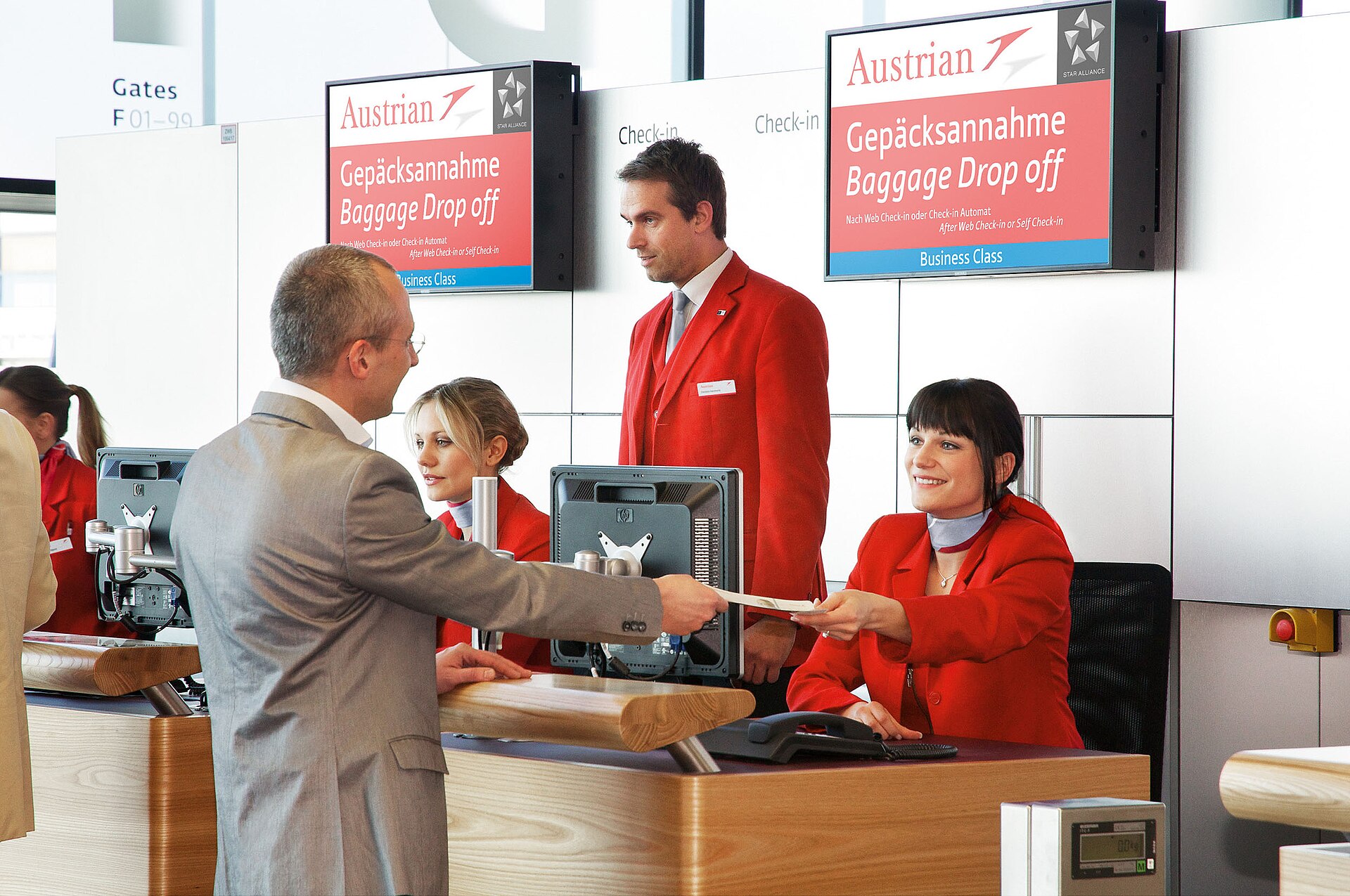 An Austrian Airlines baggage drop-off desk at Vienna Airport Terminal 3 with staff in red uniforms serving a passenger