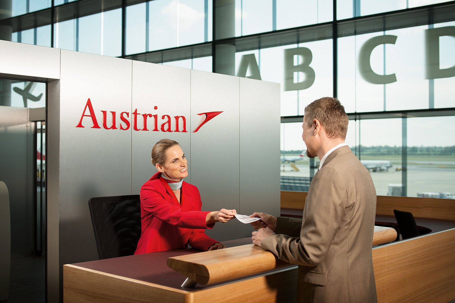 An Austrian Airlines agent in a red uniform handing a boarding pass across a check-in desk to a passenger, with large gate letters A B C D on the glass wall behind