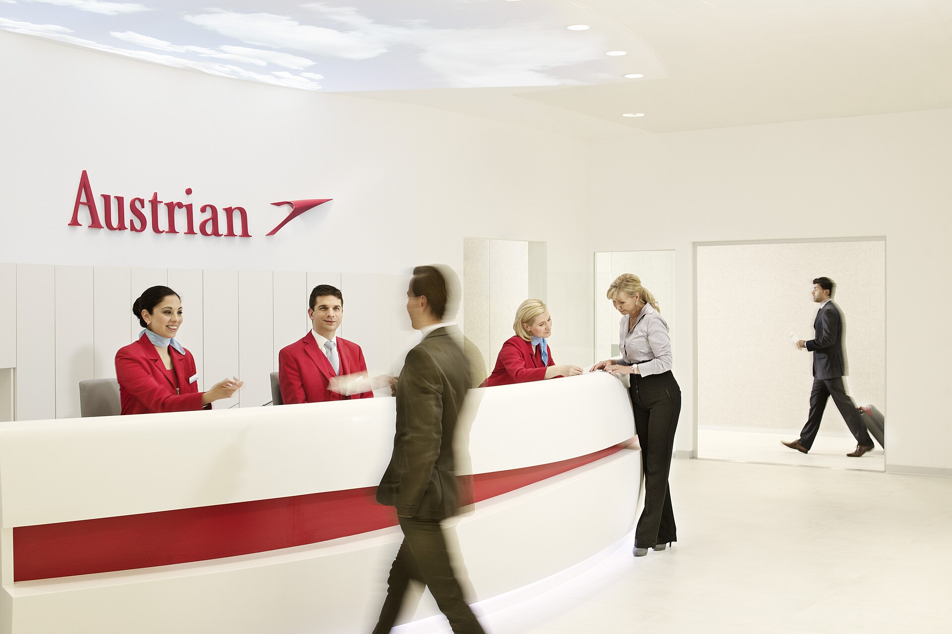 Austrian Airlines lounge reception desk at Vienna Airport with three staff in red uniforms checking in passengers under the Austrian logo