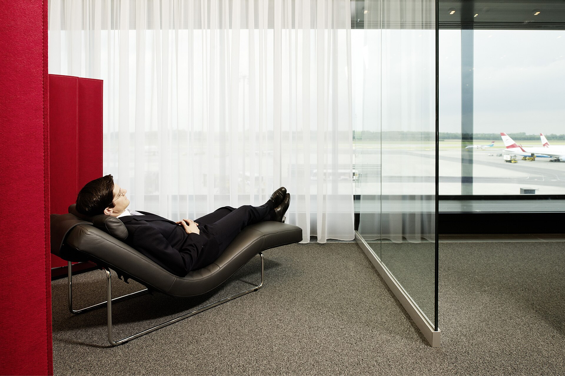 A passenger in a suit reclining on a leather chaise longue in a quiet zone of an Austrian Airlines lounge, with red felt screens and a view of Austrian aircraft on the apron