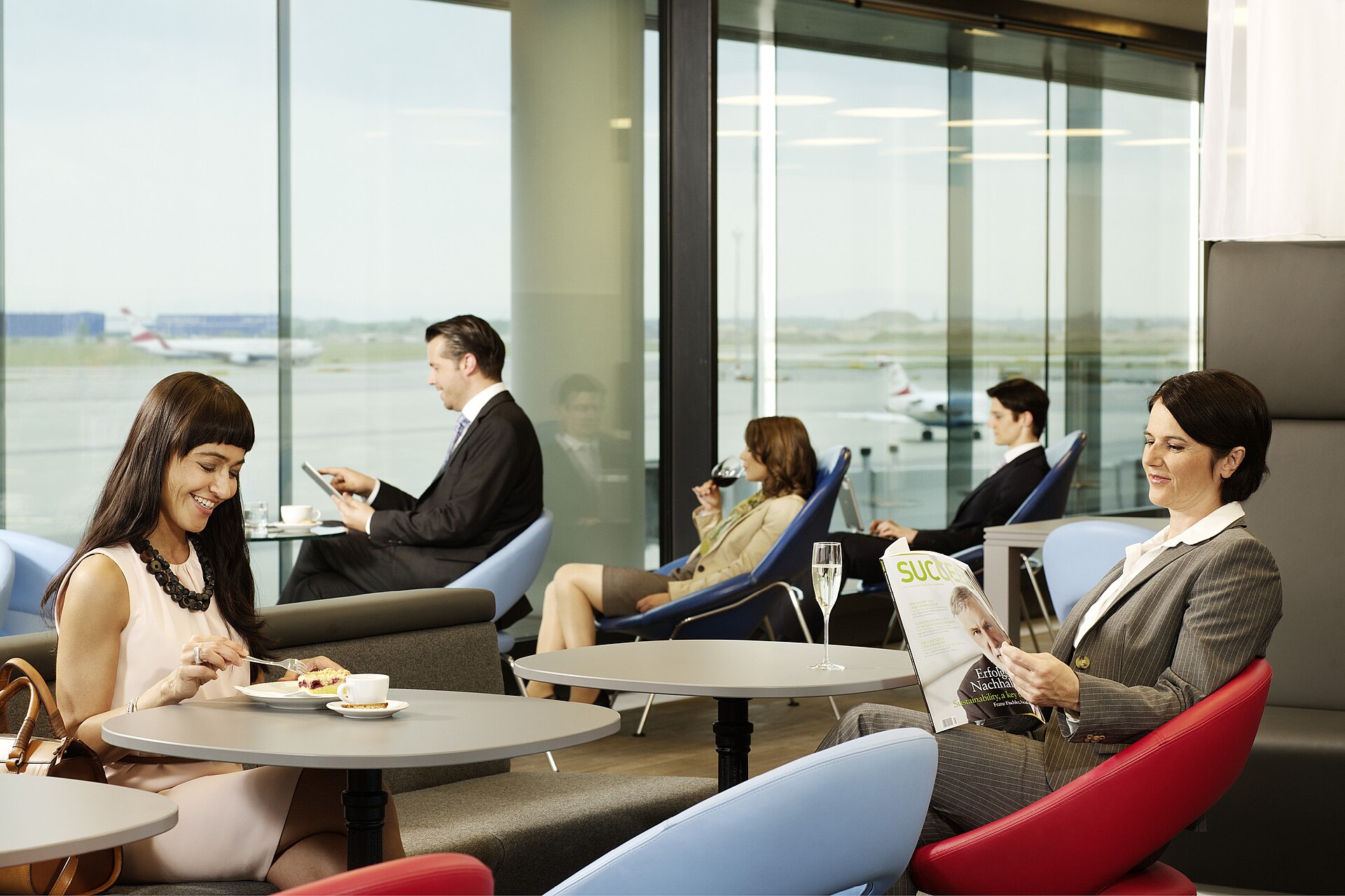 Travellers eating cake and reading in an Austrian Airlines lounge at Vienna Airport, with floor-to-ceiling windows onto the apron and a red-tailed Austrian jet outside