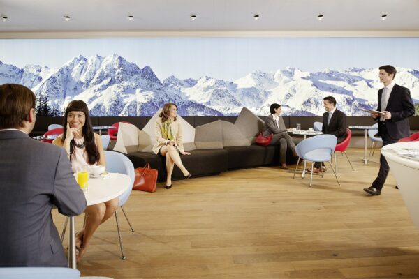 Travellers relaxing in an Austrian Airlines lounge at Vienna Airport beneath a wall-length backlit panorama of snow-covered Alpine peaks