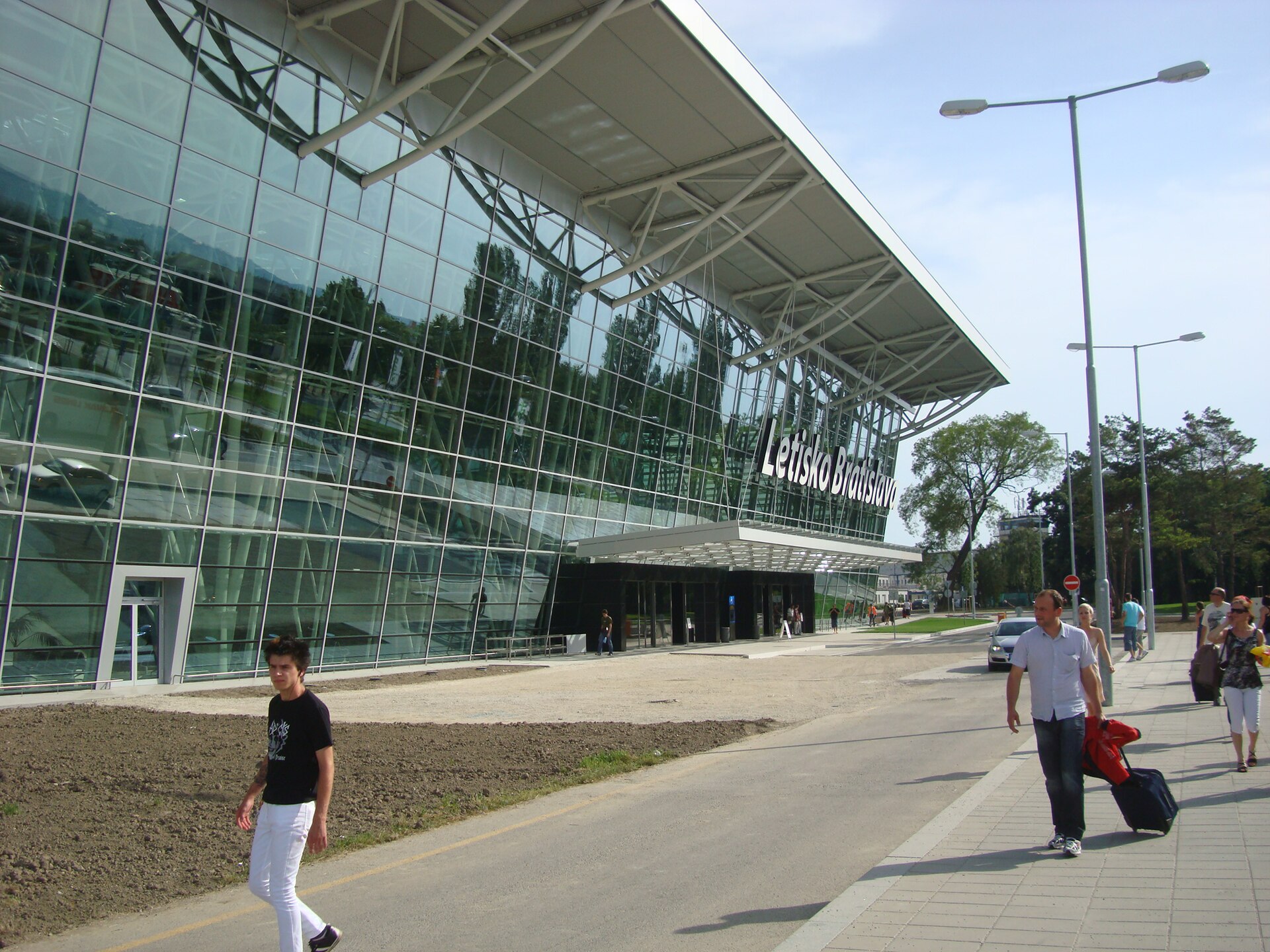 The glass facade of the passenger terminal at Bratislava Airport with the sign Letisko Bratislava and travellers wheeling suitcases along the pavement