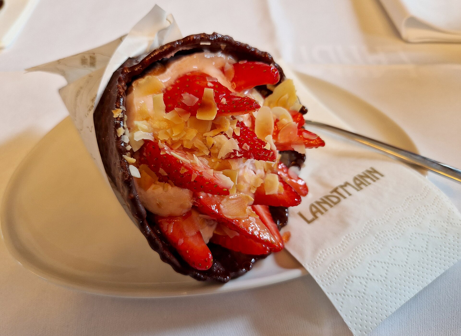 A chocolate-lined pastry horn filled with strawberry cream, sliced fresh strawberries and toasted flaked almonds, on a white plate with a Landtmann napkin