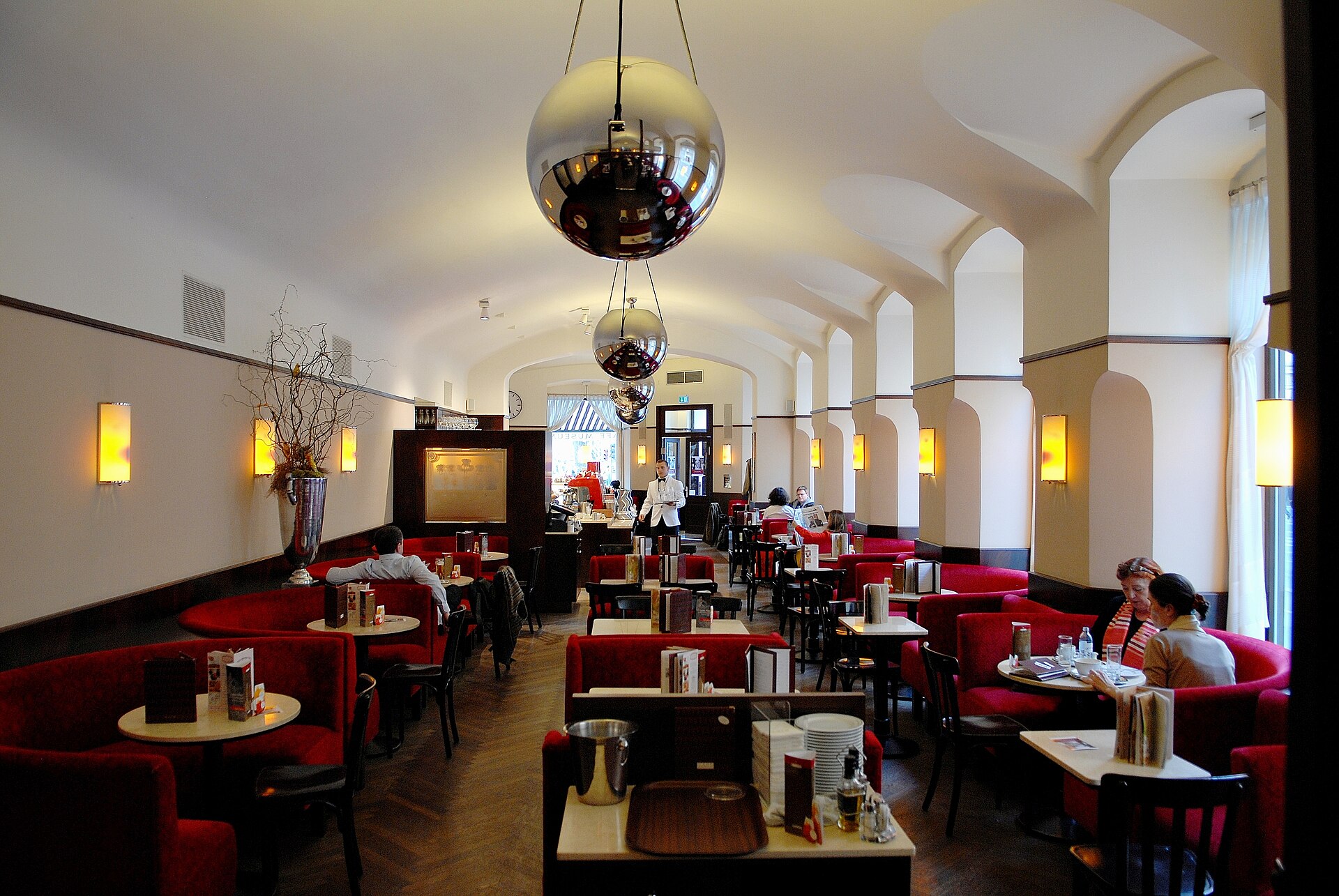 The long interior of Café Museum in Vienna, with red leather banquettes and chairs, small tables, arched windows along one side, wall lamps and large mirrored globe pendant lights