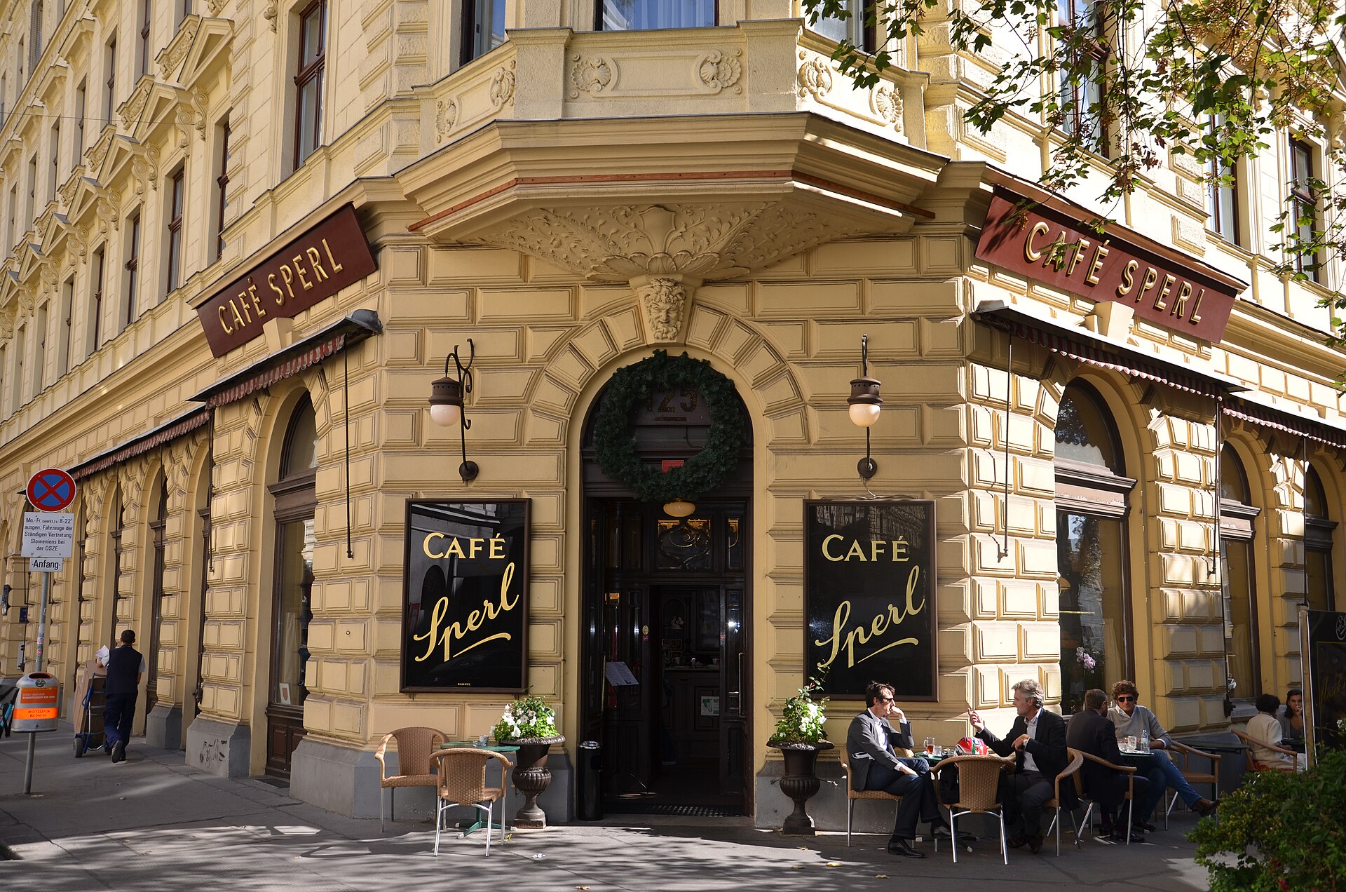 The corner entrance of Café Sperl on Gumpendorfer Straße in Vienna, with a pale yellow facade, gold script lettering on black signs reading Café Sperl and guests at outdoor tables
