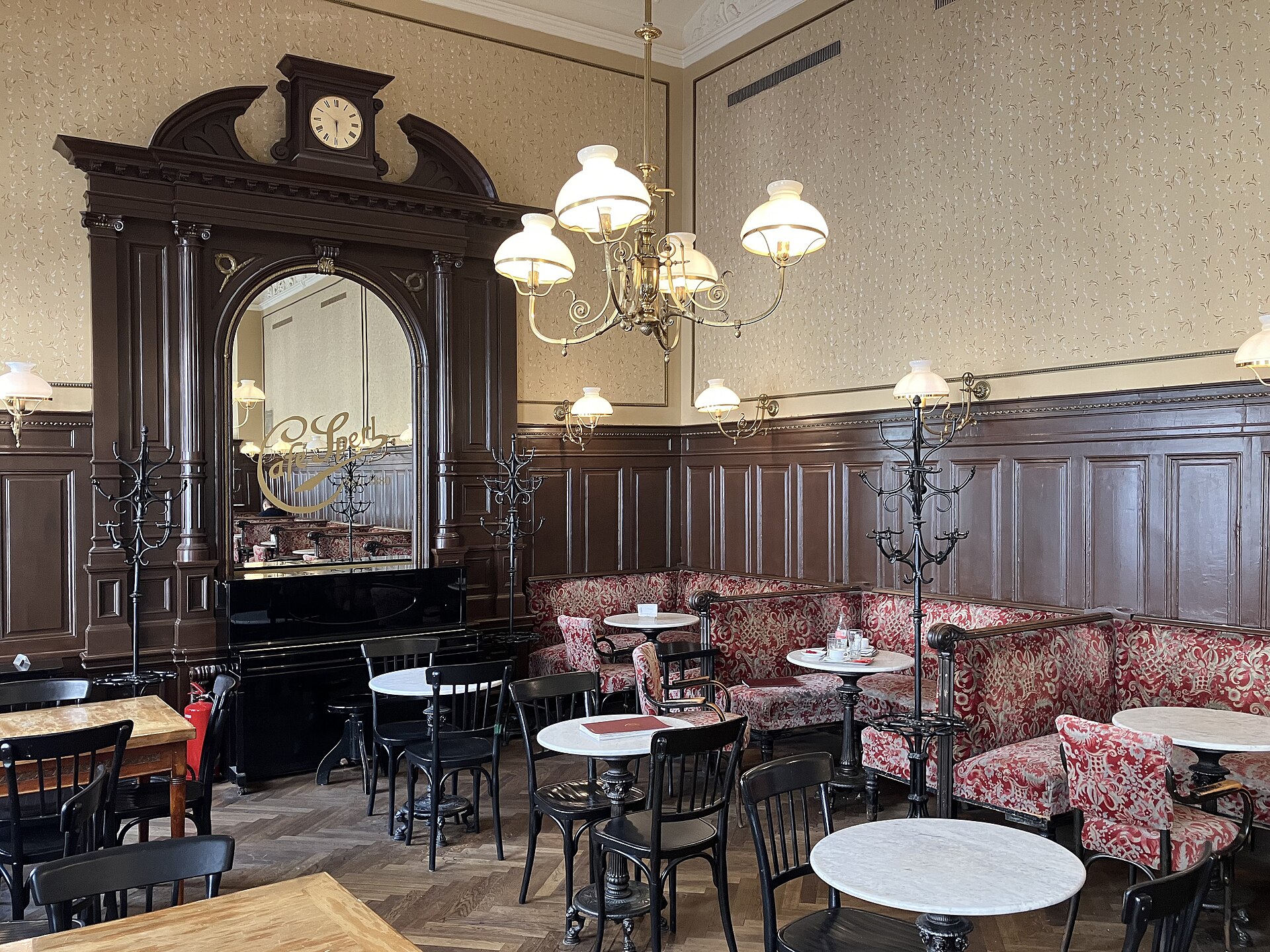 The interior of Café Sperl in Vienna, with a tall carved dark wood cabinet and mirror, a clock, dark panelled walls, red patterned upholstered banquettes, white marble tables and chandeliers