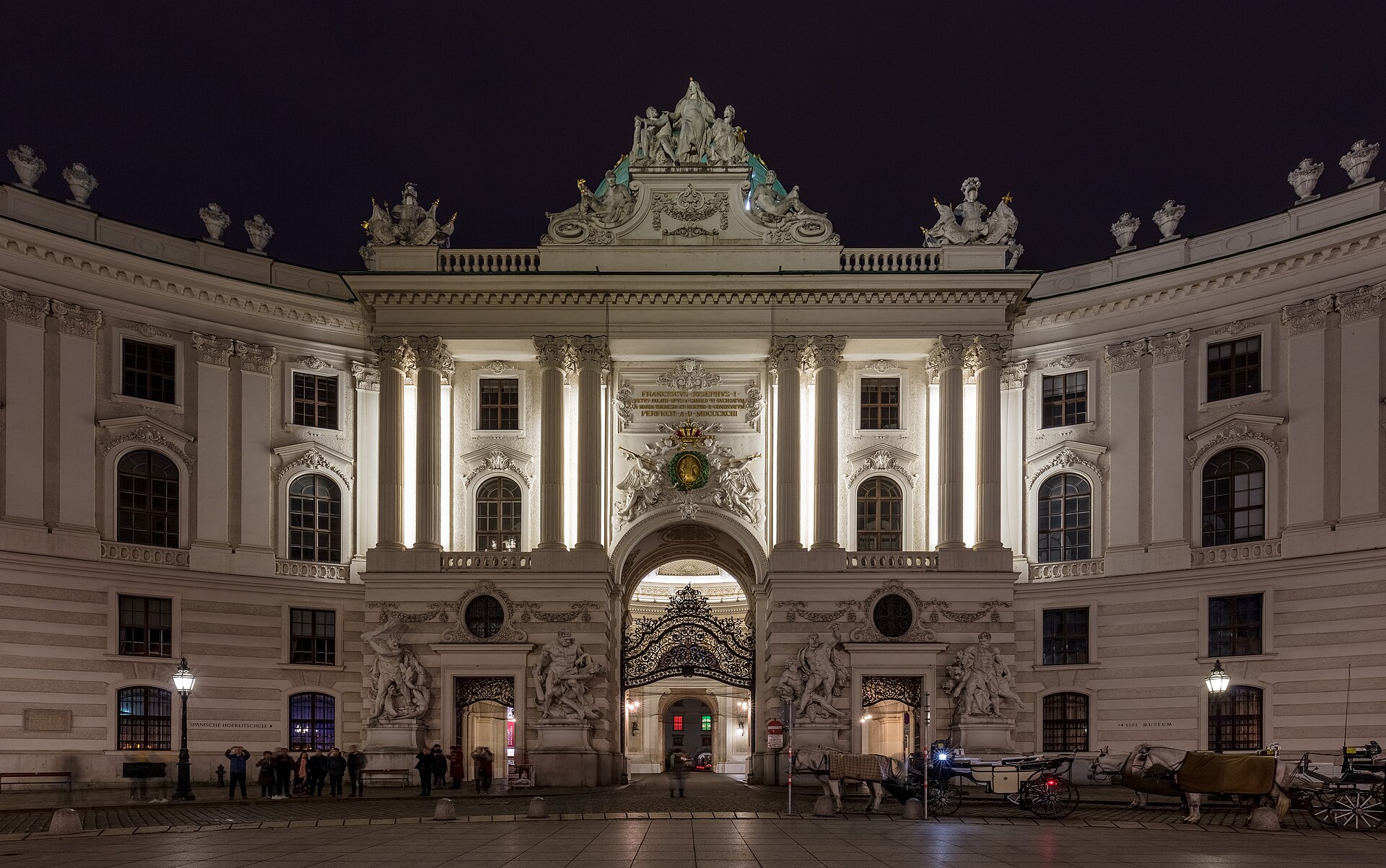 The floodlit Michaelertrakt of the Hofburg at night, its domed gateway lit from below, with fiaker carriages waiting on the cobbles and a lit Sisi Museum sign on the right-hand wing