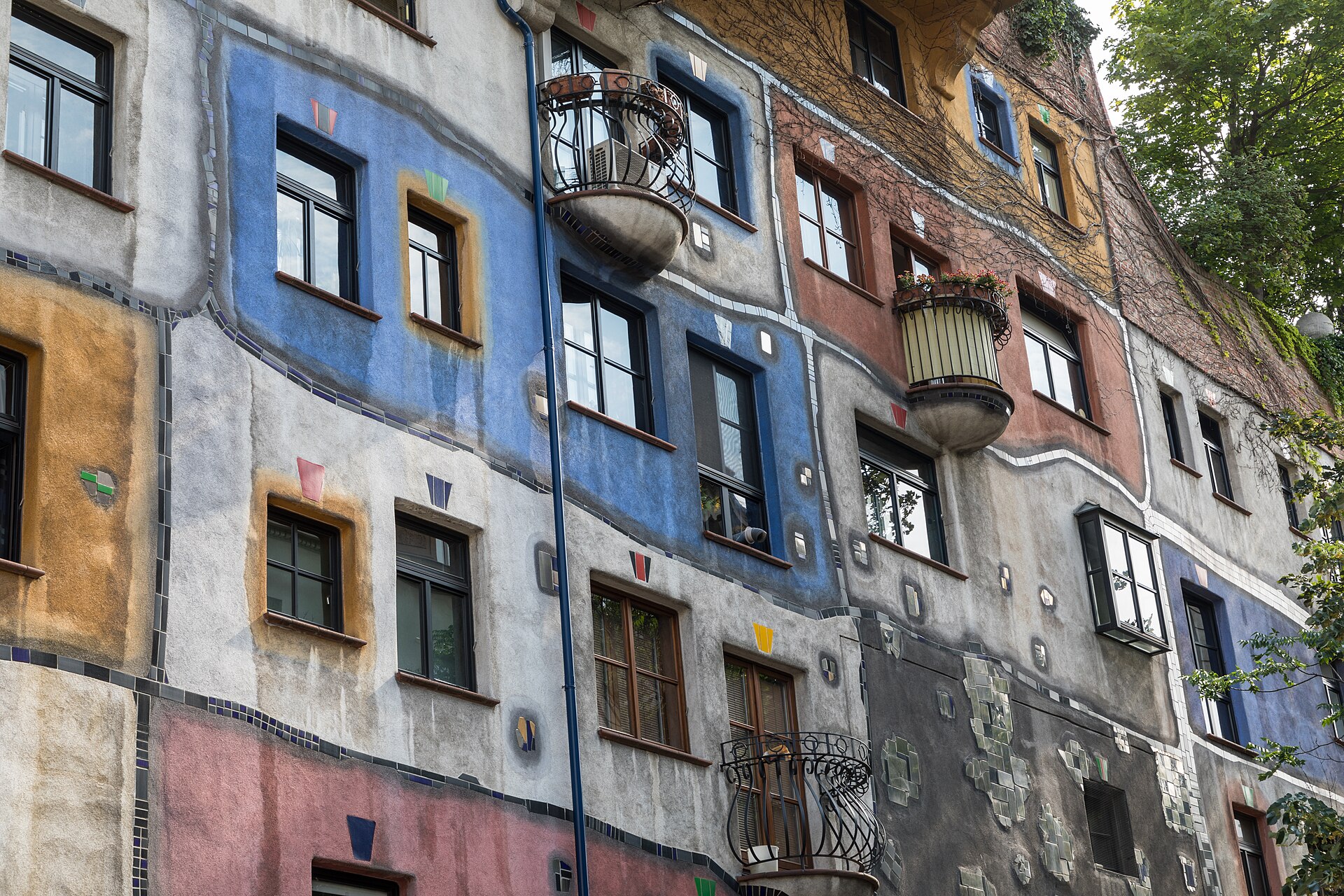 The Hundertwasserhaus in Vienna with its patchwork facade of blue, pink and white panels, irregular windows, black balcony railings and plants growing from the balconies