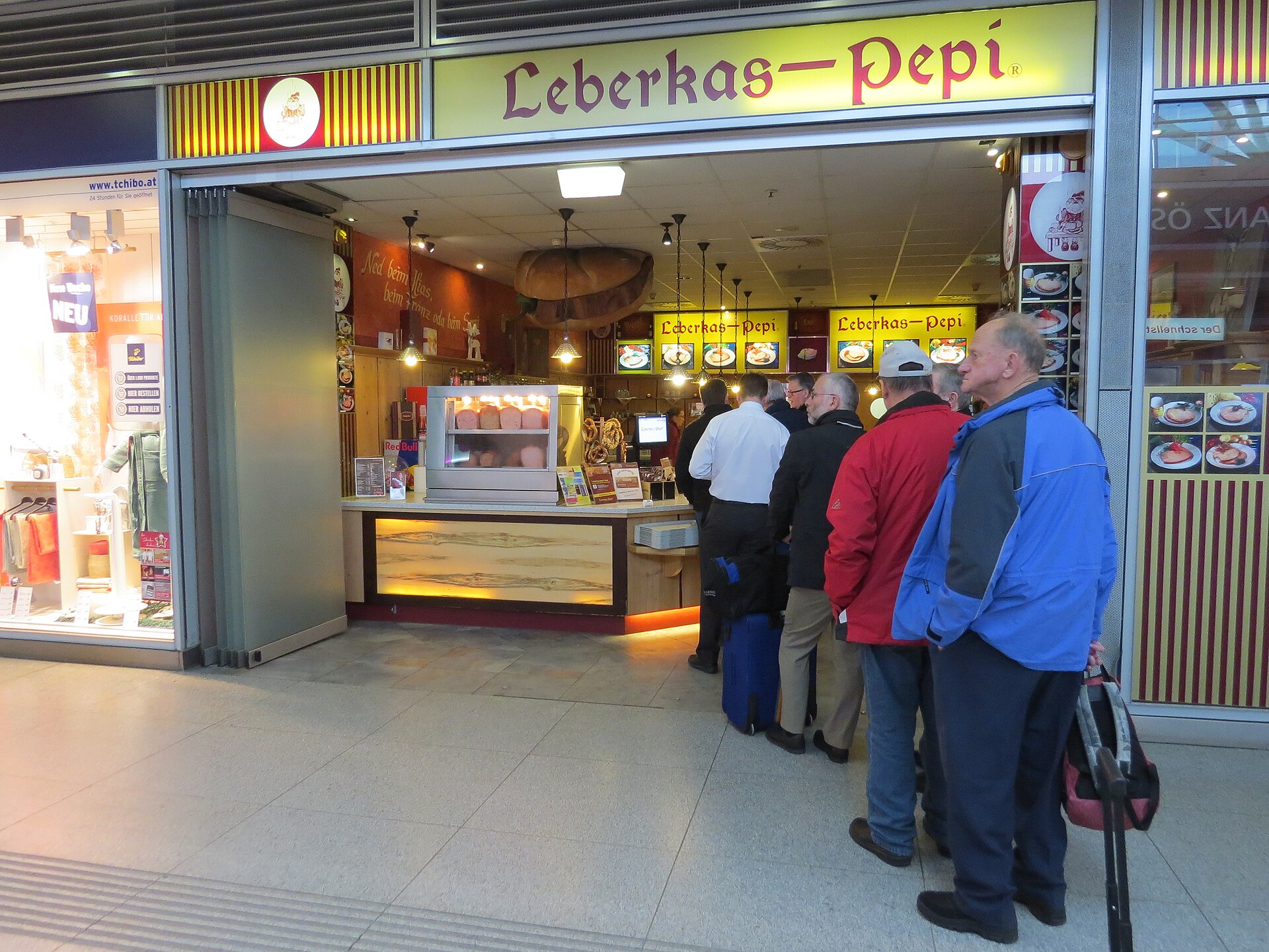 Travellers with luggage queuing at a Leberkas-Pepi counter in a station concourse, with loaves of Leberkäse in a heated display and a yellow illuminated sign