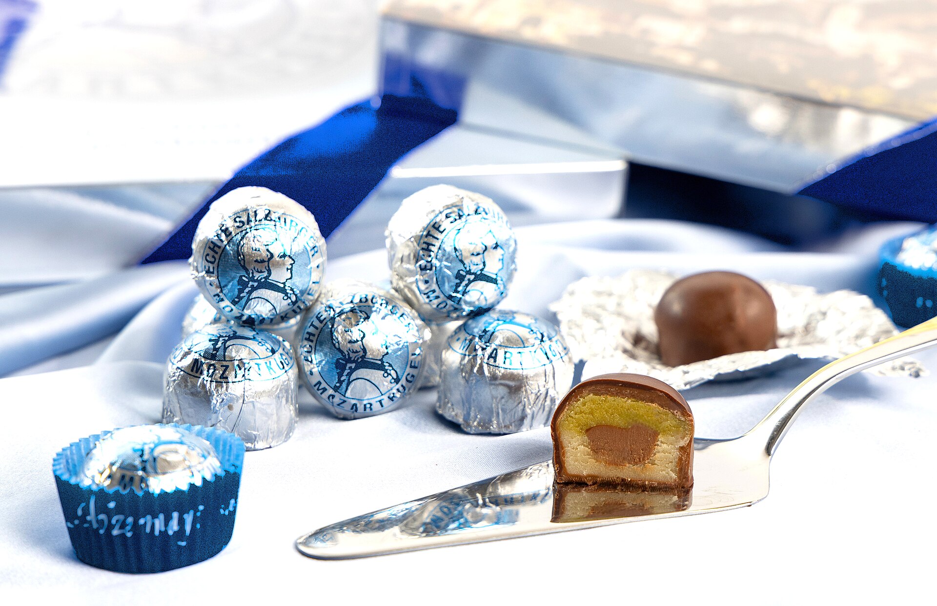 Foil-wrapped Mozartkugeln printed with a portrait of Mozart beside one chocolate cut in half to show green pistachio marzipan and a nougat centre on a silver cake server