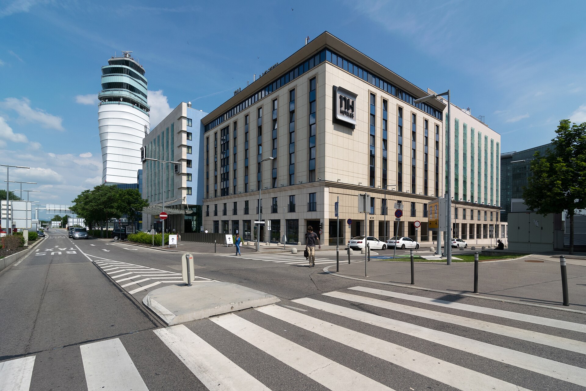 The NH Hotel at Vienna Airport, a pale eight-storey building on a street corner with a pedestrian crossing, with the white airport control tower rising behind it