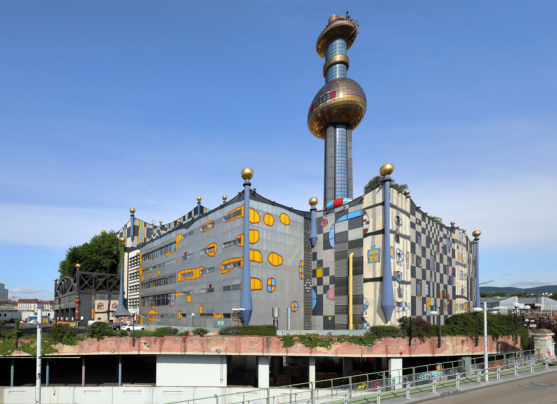 The Spittelau waste incineration plant in Vienna with its gold-clad sphere on a tall chimney, chequerboard and mosaic facade panels and blue roof details