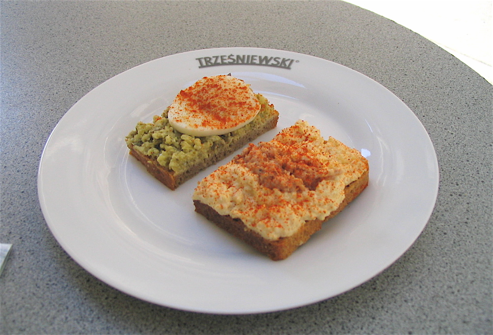 Two small open sandwiches on dark bread on a white Trzesniewski plate, one topped with egg spread and a slice of boiled egg, the other with a creamy spread dusted with paprika