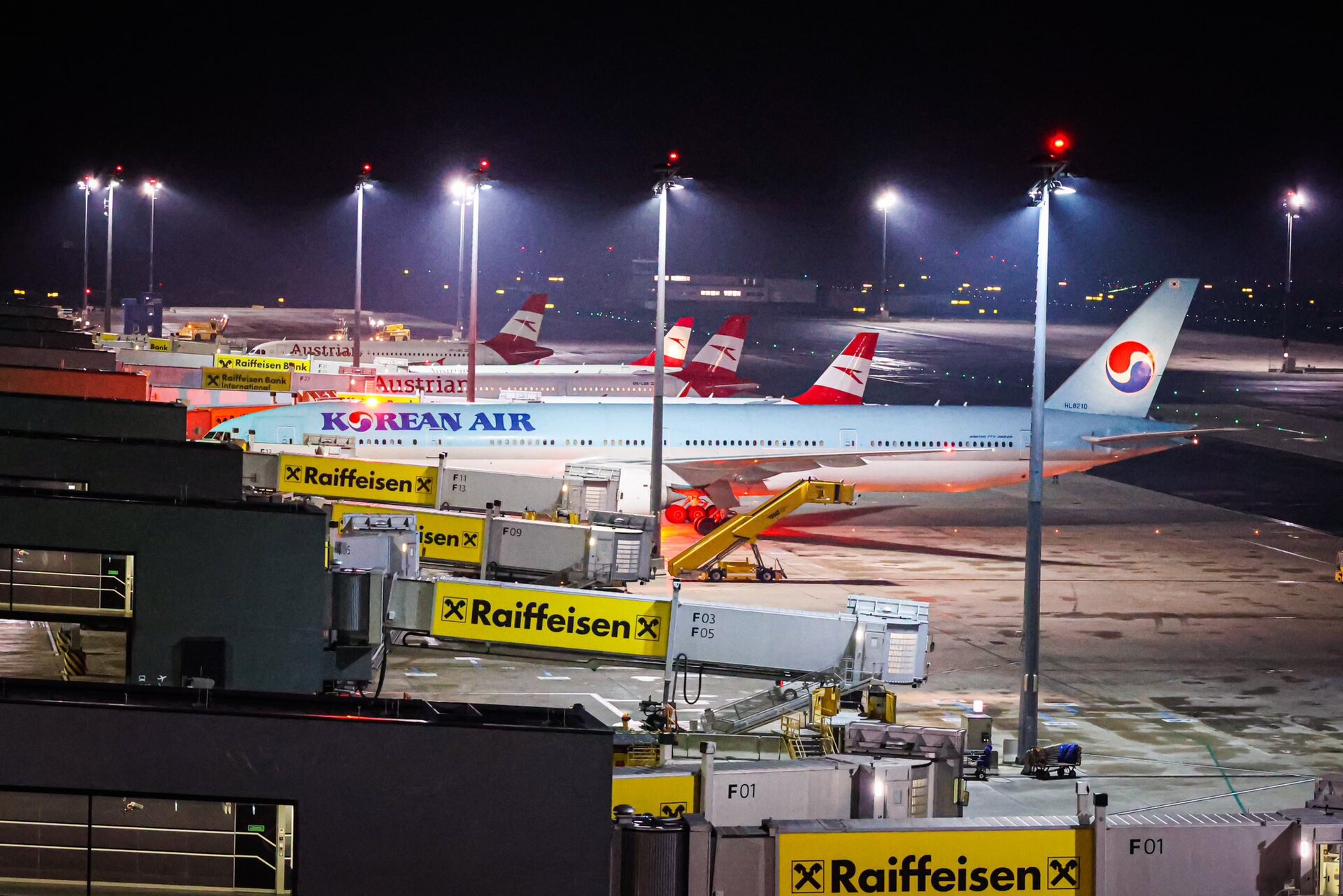 Austrian Airlines and Korean Air aircraft parked at the Pier North jet bridges at Vienna Airport at night under floodlights, with gate numbers F01 to F13 on the bridges