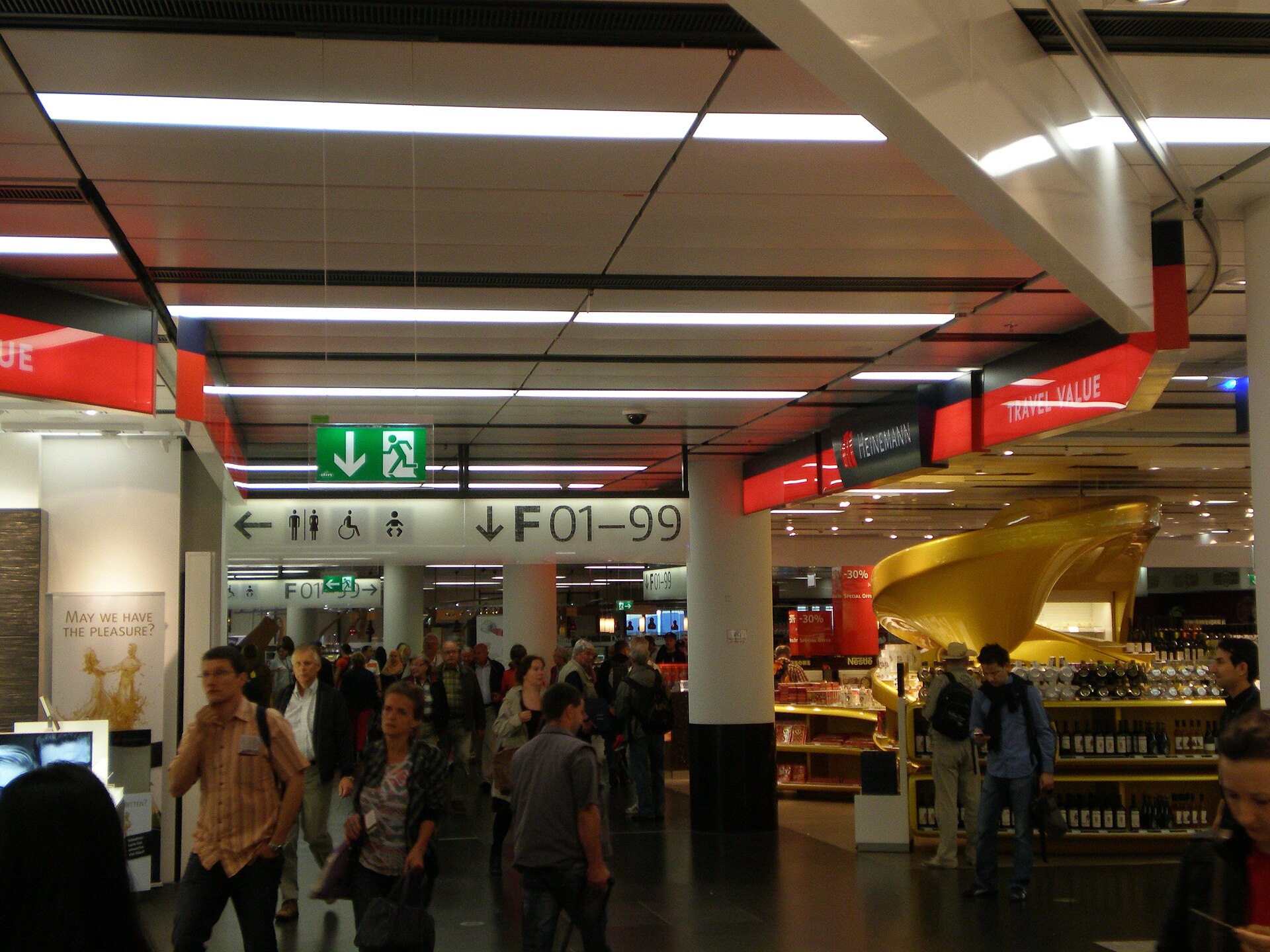 Passengers walking past the Heinemann shop by the F gates at Vienna Airport, with red Travel Value signage, shelves of wine and a large golden display stand