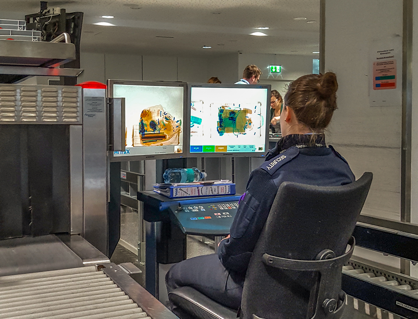 A security officer at Vienna Airport watching two monitors showing X-ray images of hand luggage beside a baggage screening machine
