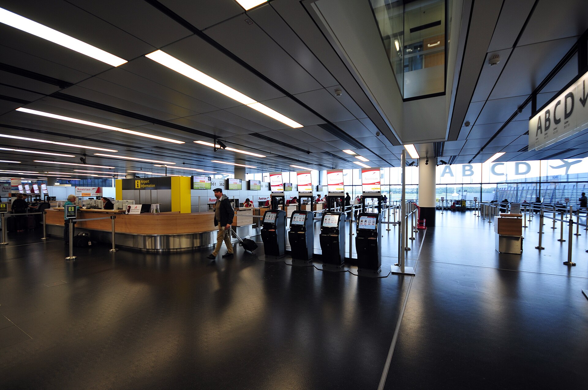 The Terminal 3 check-in hall at Vienna Airport with self-service kiosks and large A B C D gate signage above the entrance to the piers