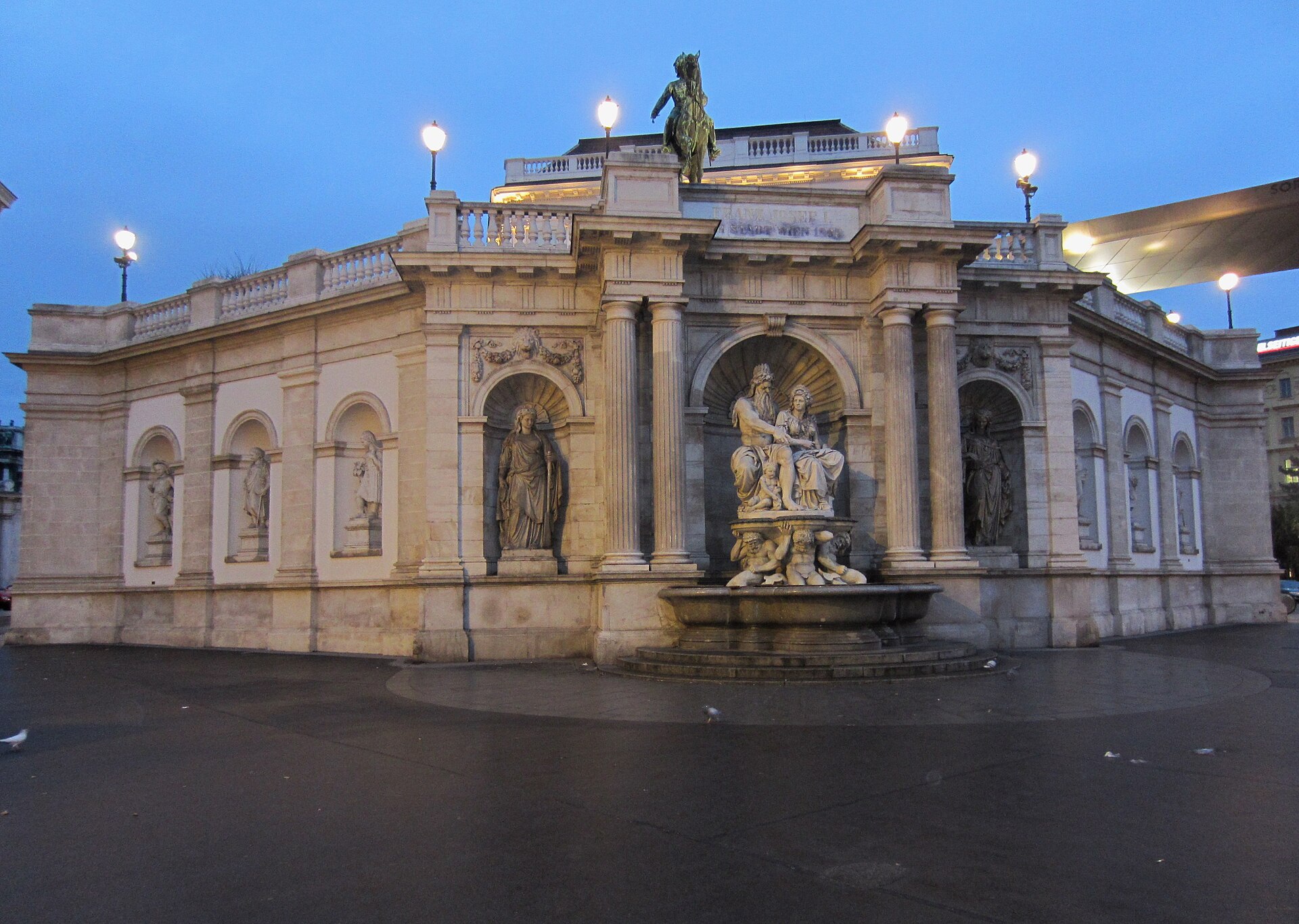The Albrechtsbrunnen fountain at the street-level base of the Albertina in Vienna at dusk, with the equestrian statue of Archduke Albrecht on the terrace above