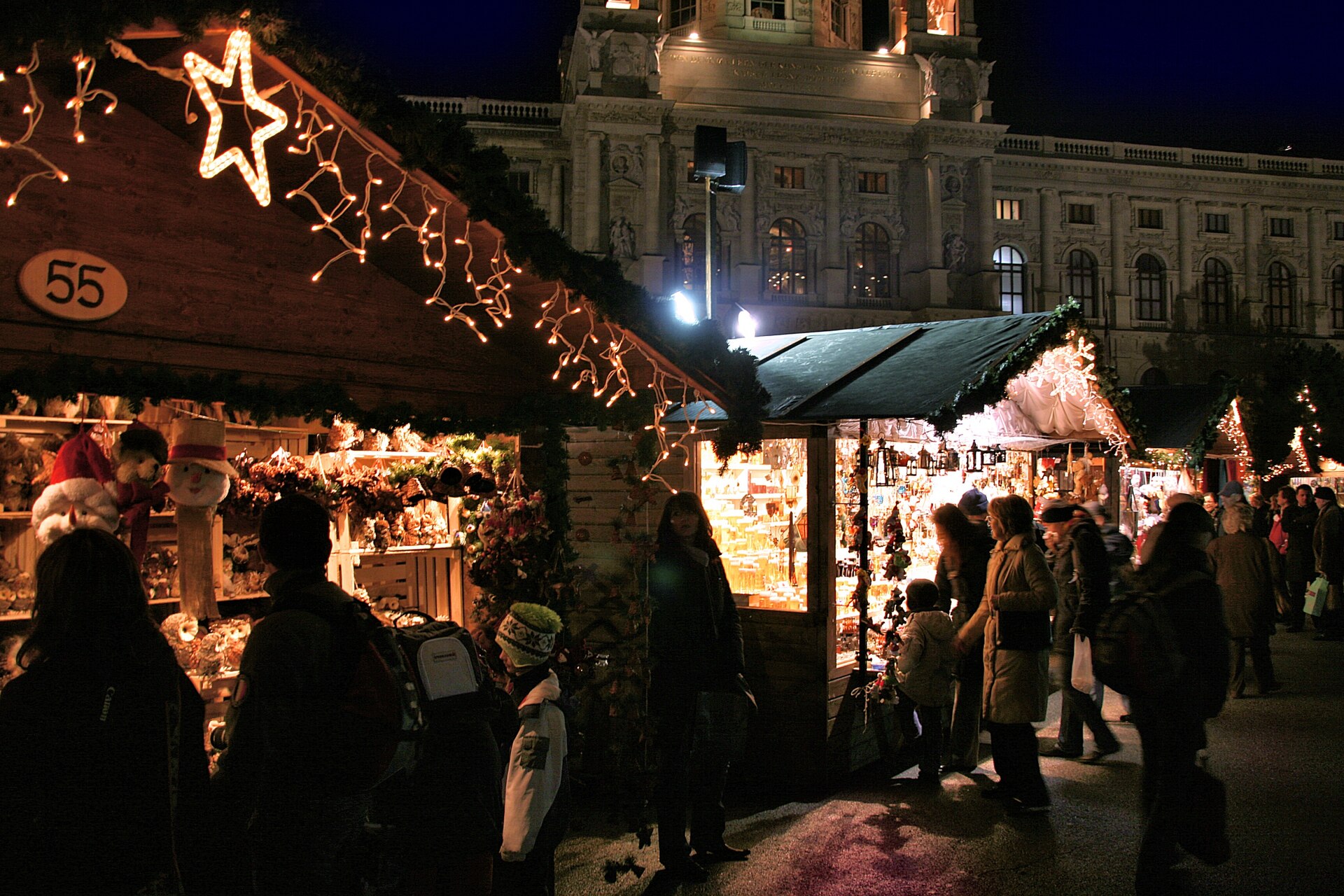 Illuminated wooden Christmas market stalls hung with lights and decorations at night in Vienna, with a floodlit museum facade behind