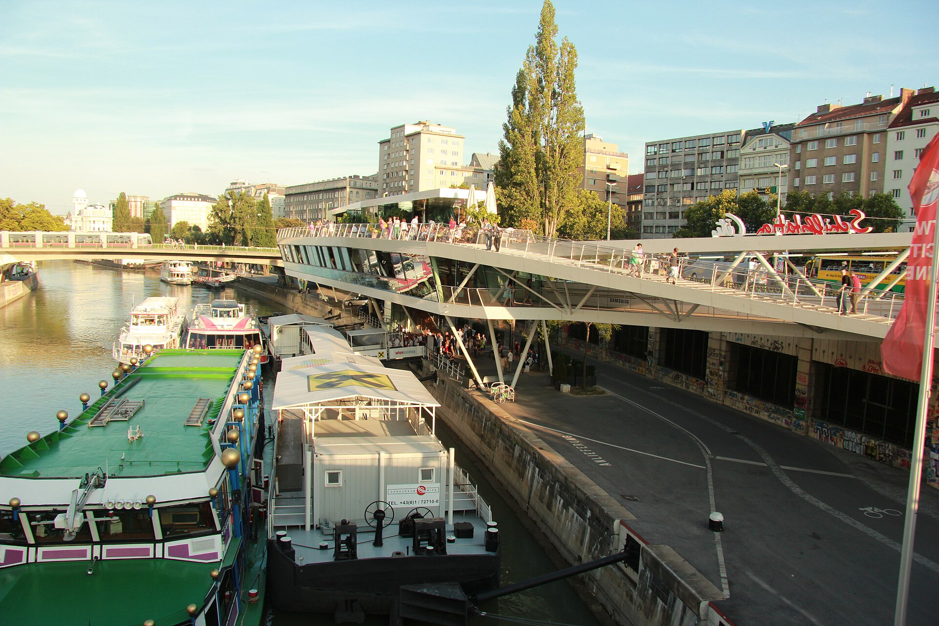 Boats moored along the Donaukanal at the Schwedenplatz shipping station in Vienna, with the curved glass Schiffstation building and a footbridge above