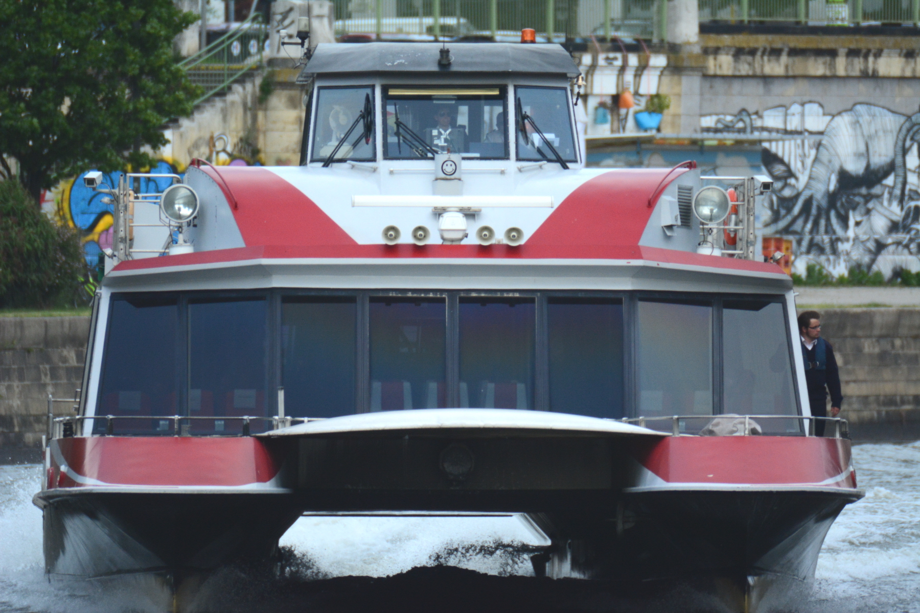 A red and white catamaran seen head-on travelling fast along the Donaukanal in Vienna with graffitied embankment walls behind