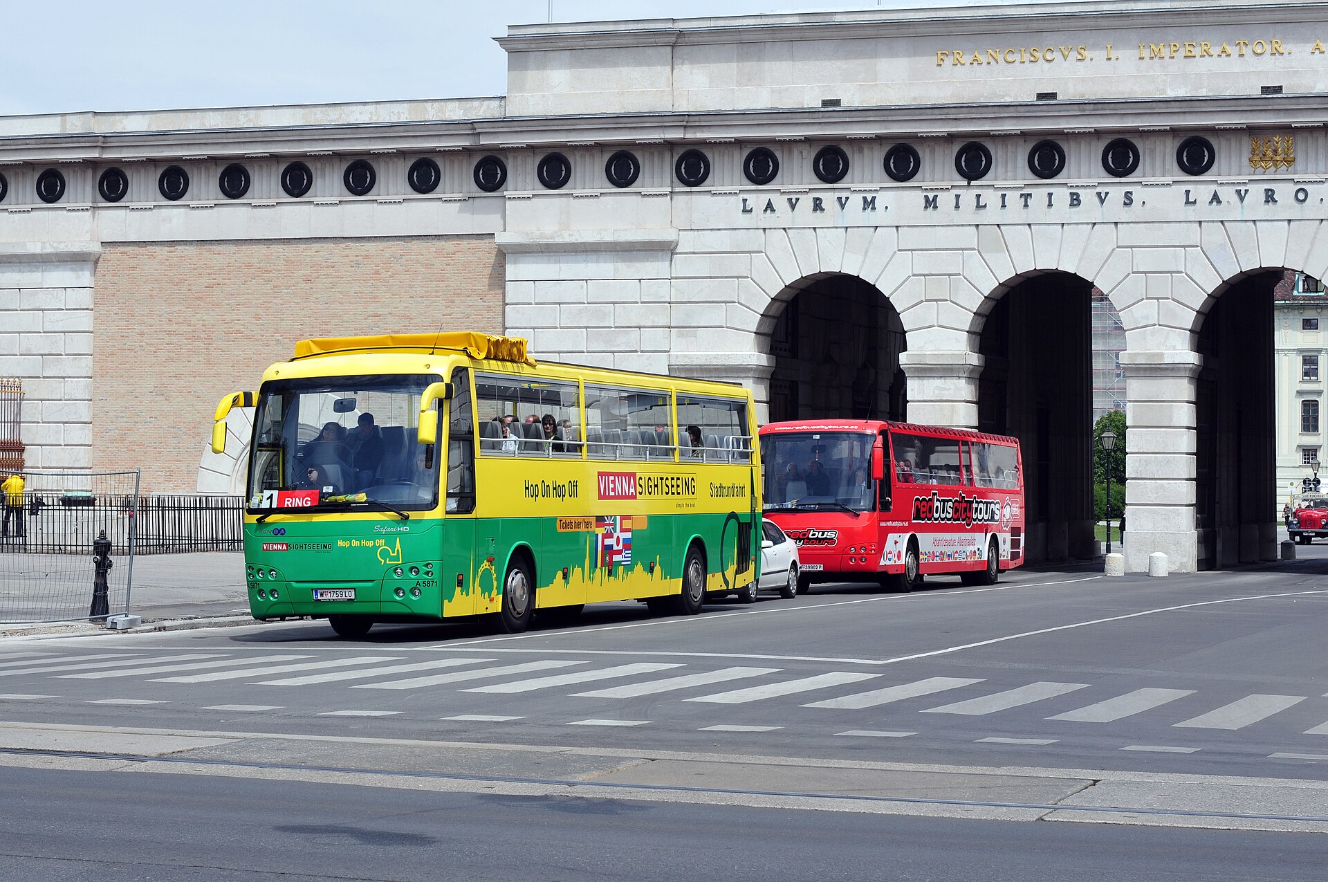 A yellow and green Vienna Sightseeing hop-on hop-off bus marked Route 1 Ring waiting beside a red Redbus City Tours coach at the Burgtor gateway in Vienna
