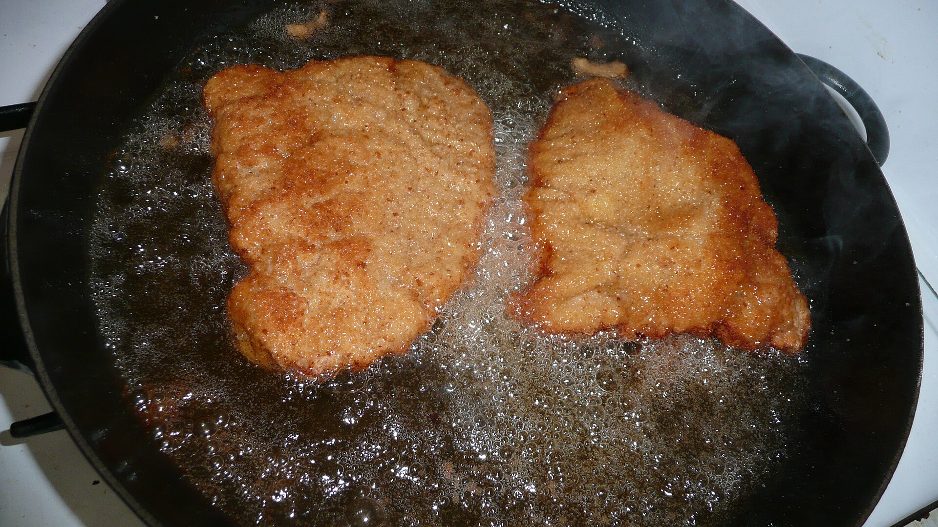 Two Wiener schnitzels frying in a large black pan with hot fat bubbling around them