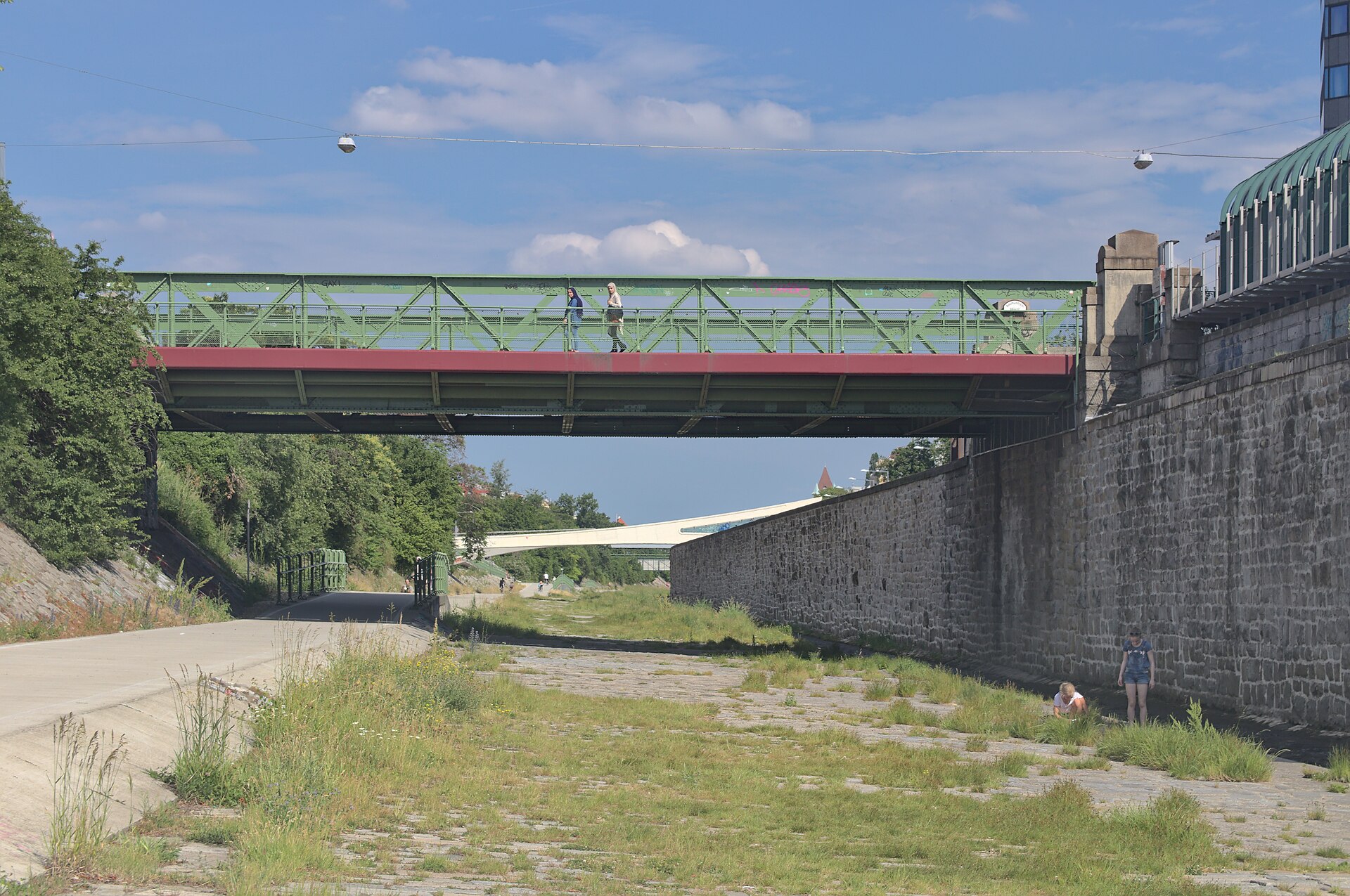 A cycle path running along the concreted Wienfluss channel beneath a green iron bridge in western Vienna