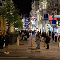 People stroll along Vienna's Kärntner Straße at night, passing illuminated shopfronts, outdoor tables, trees and bollards.